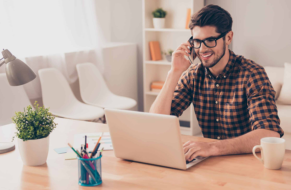 En ung, glad mann med briller som sitter i en telefonsamtale mens han sitter foran PC'en sin.