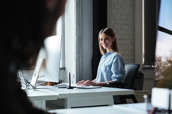 En jente som sitter på sitt skrivebord og jobber mens hun smiler til sin kollega.